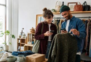 Two shoppers using secondhand shopping apps in a UK vintage clothing shop